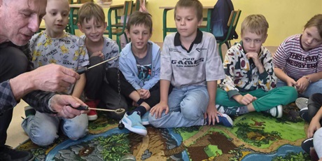 Powiększ grafikę: dzieci siedzą na dywanie i obserwują doświadczenia wykonywane przez pana chemika