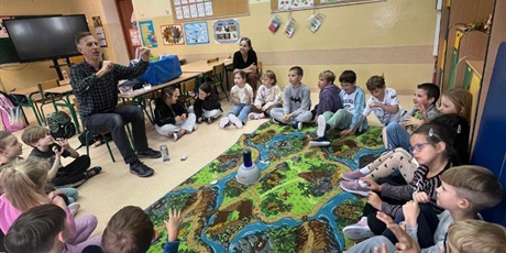 Powiększ grafikę: dzieci siedzą na dywanie i obserwują doświadczenia wykonywane przez pana chemika