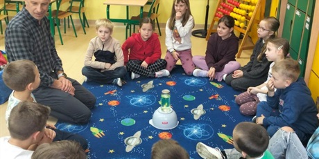 Powiększ grafikę: dzieci siedzą na dywanie i obserwują doświadczenia wykonywane przez pana chemika